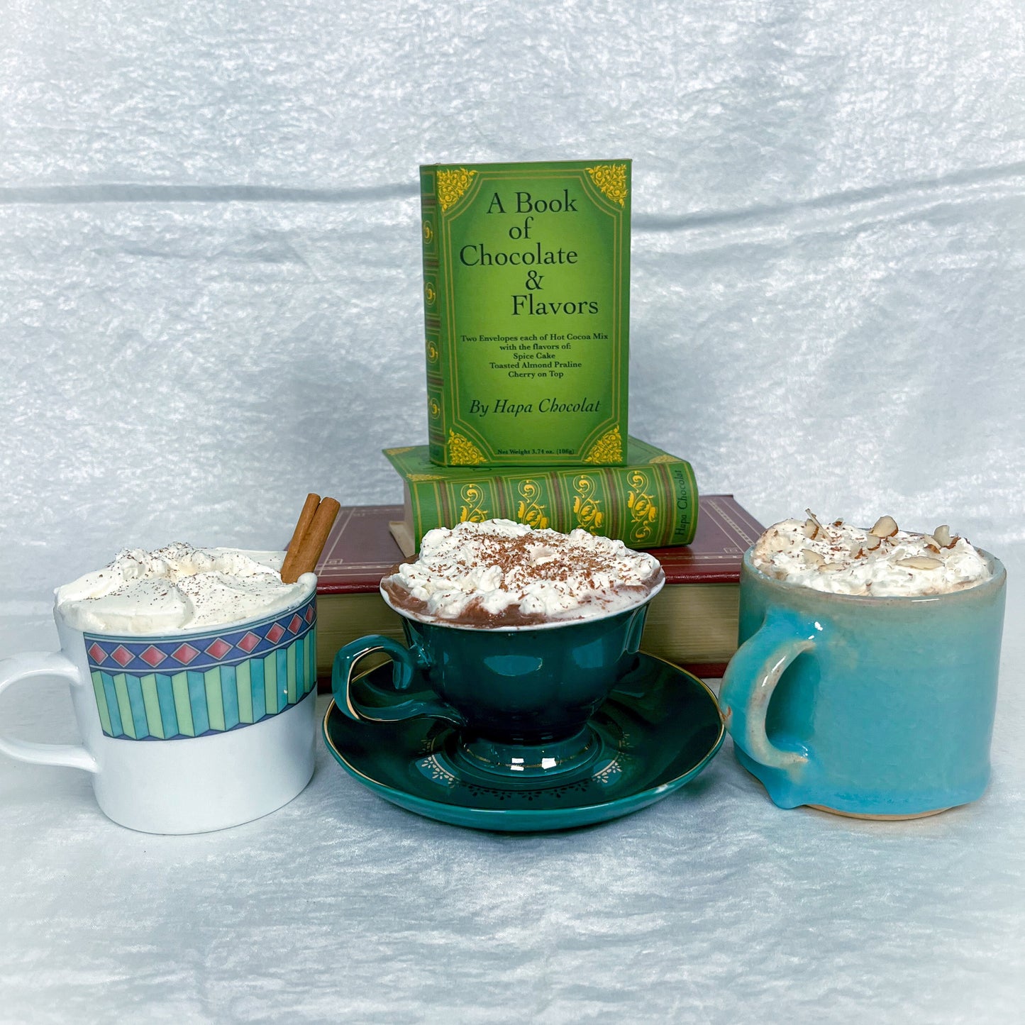 Two green books and 3 cups of hot cocoa with whipped cream in different styles of mugs.
