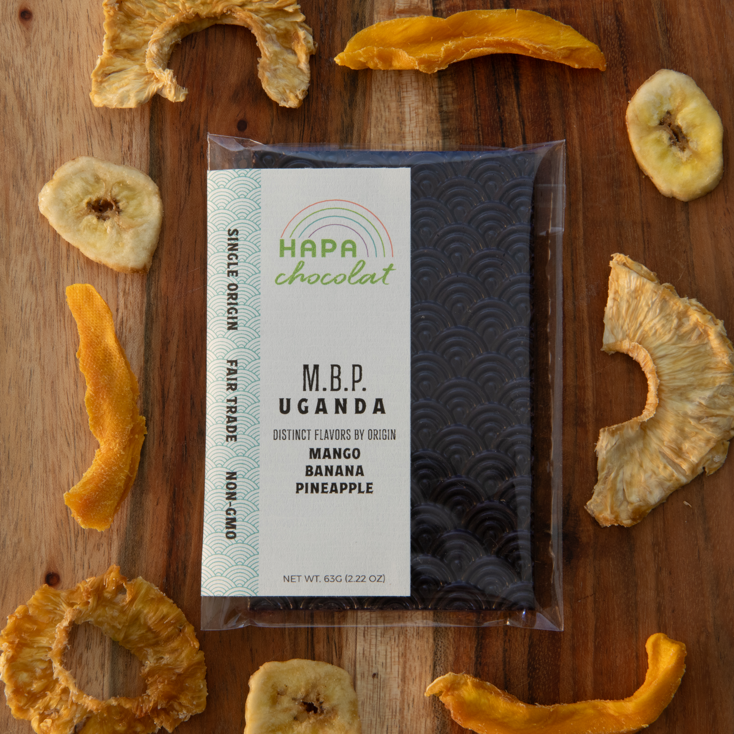 A seigaiha pattern dark chocolate bar in an eco-cello bag with an ivory colored label with the logo and product descriptions, framed by dried mango and dried pineapple pieces, and a few banana chips.