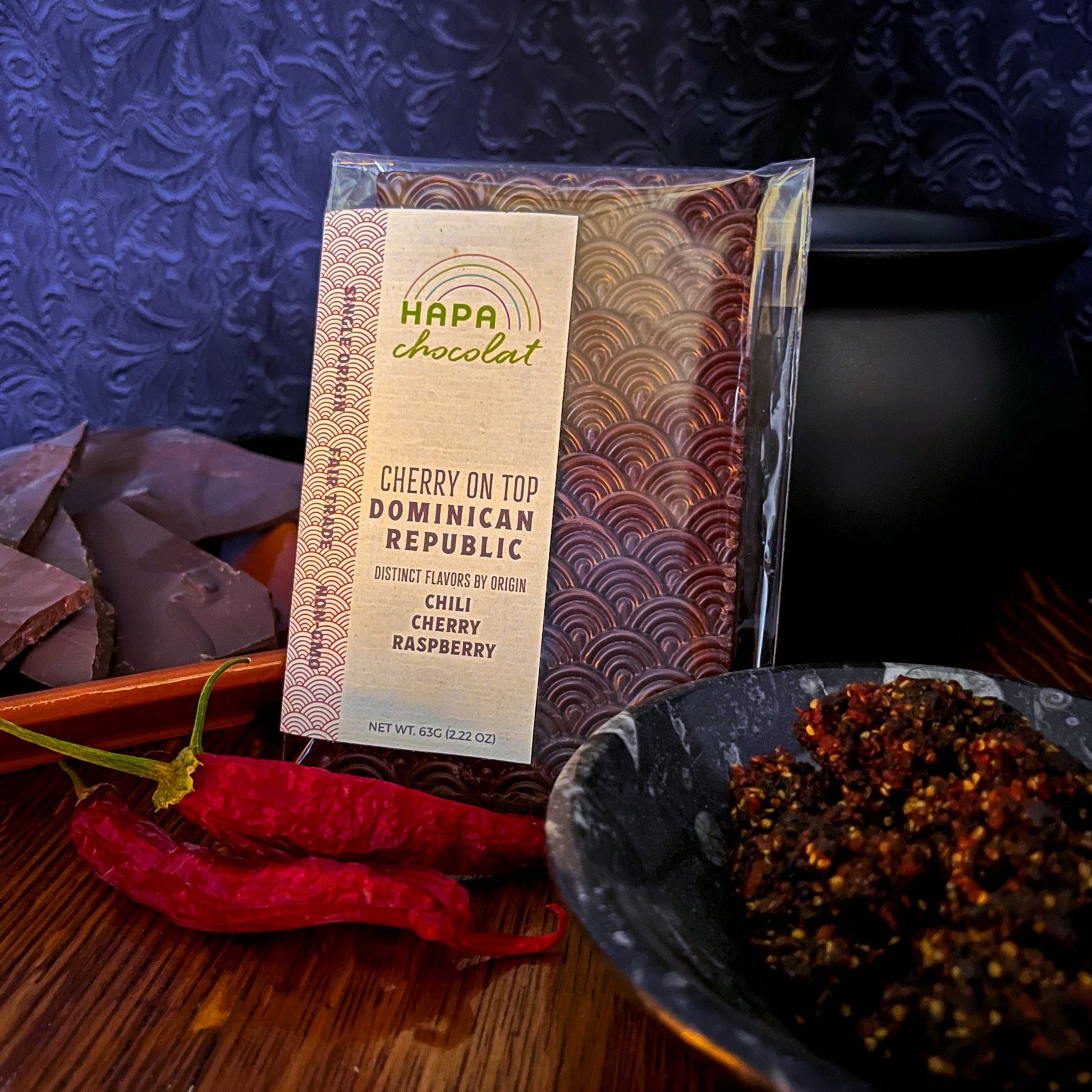 A packaged chocolate bar propped against a cauldron, in front of shards of dark chocolate, with dried chilis in front, and a bowl of crushed dried cherries. All are ontop of a wooden surface, and a swirly purple-black backdrop.