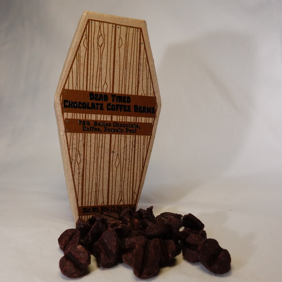 A cardboard coffin with printed wood grain has a label that reads 'Dead Tired Chocolate Coffee Beans'. Under that is another label that says '72% Belize Chocolate, Coffee, Pomelo Peel.'

At the base of the coffin are coffee bean shape chocolates.

The background is white.