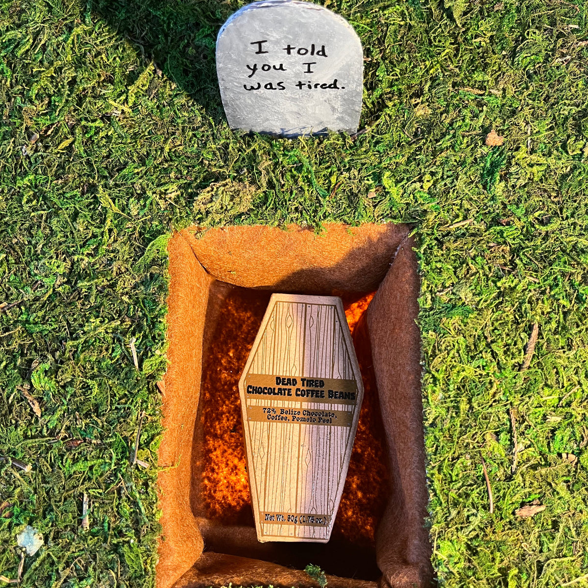 A sunset grave site with a cardboard coffin labelled 'Dead Tired Chocolate Coffee Beans' laying in the grave. Above it is a tombstone that reads 'I told you I was tired.'