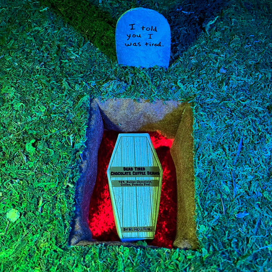 A nighttime grave site with a cardboard coffin labelled 'Dead Tired Chocolate Coffee Beans' laying in the grave that is lit red. Above it is a tombstone that reads 'I told you I was tired.'