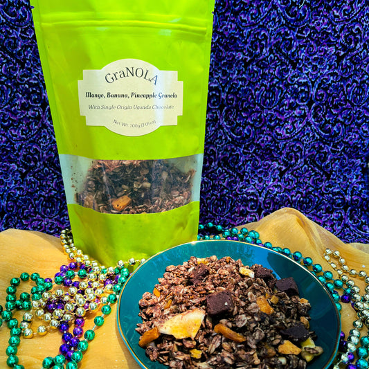 A bright green full of chocolate covered granola is in front of a purple and black swirled background. In the foreground is a dark green bow full of the granola with chunks of chocolate, banana chips, mango, and pineapple in it. All of it sits on a gold fabric with Mardi Gras beads scattered about.