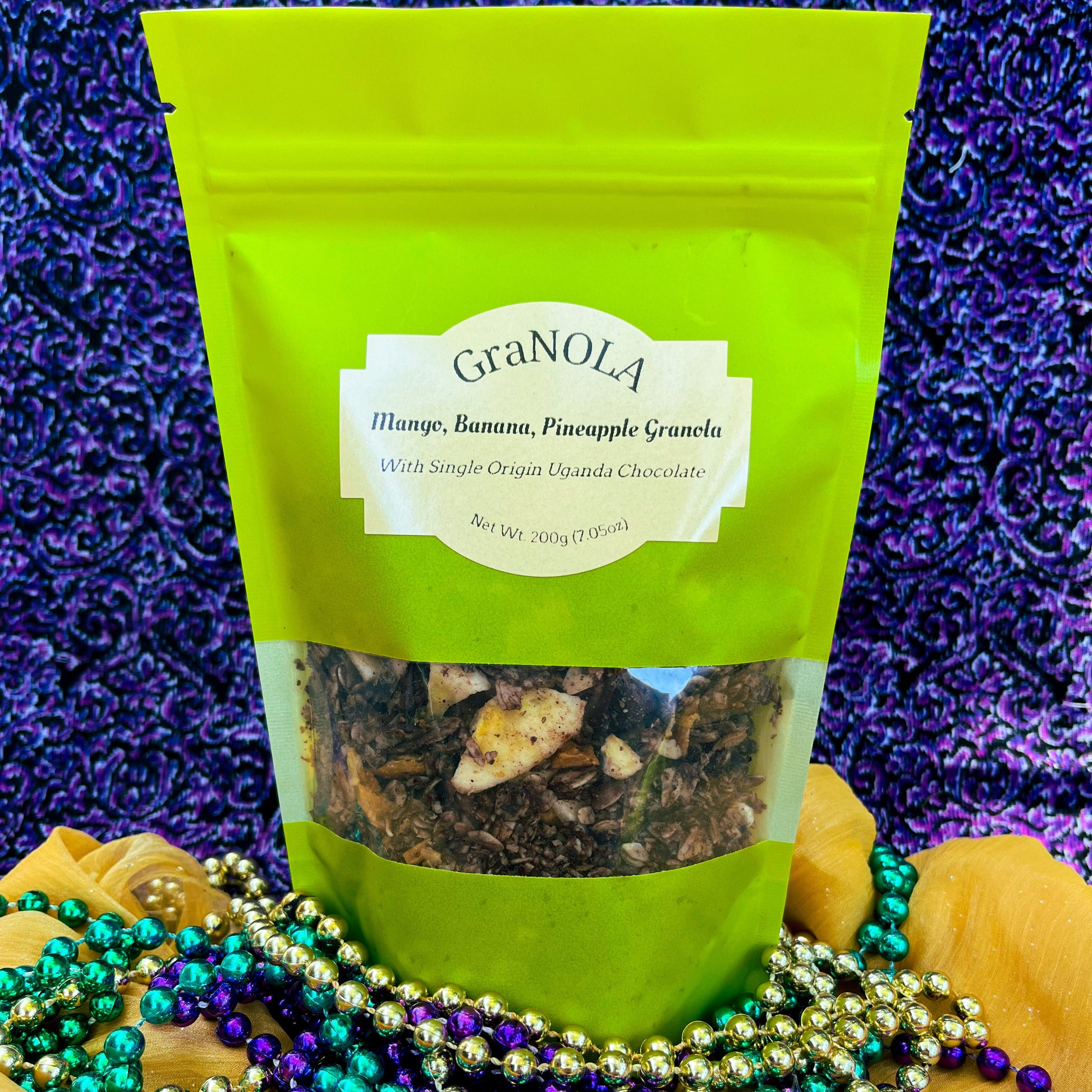 A bright green full of chocolate covered granola with chunks of chocolate, banana chips, mango, and pineapple in it. In front of a purple and black swirled background. All of it sits on a gold fabric with Mardi Gras beads scattered about.