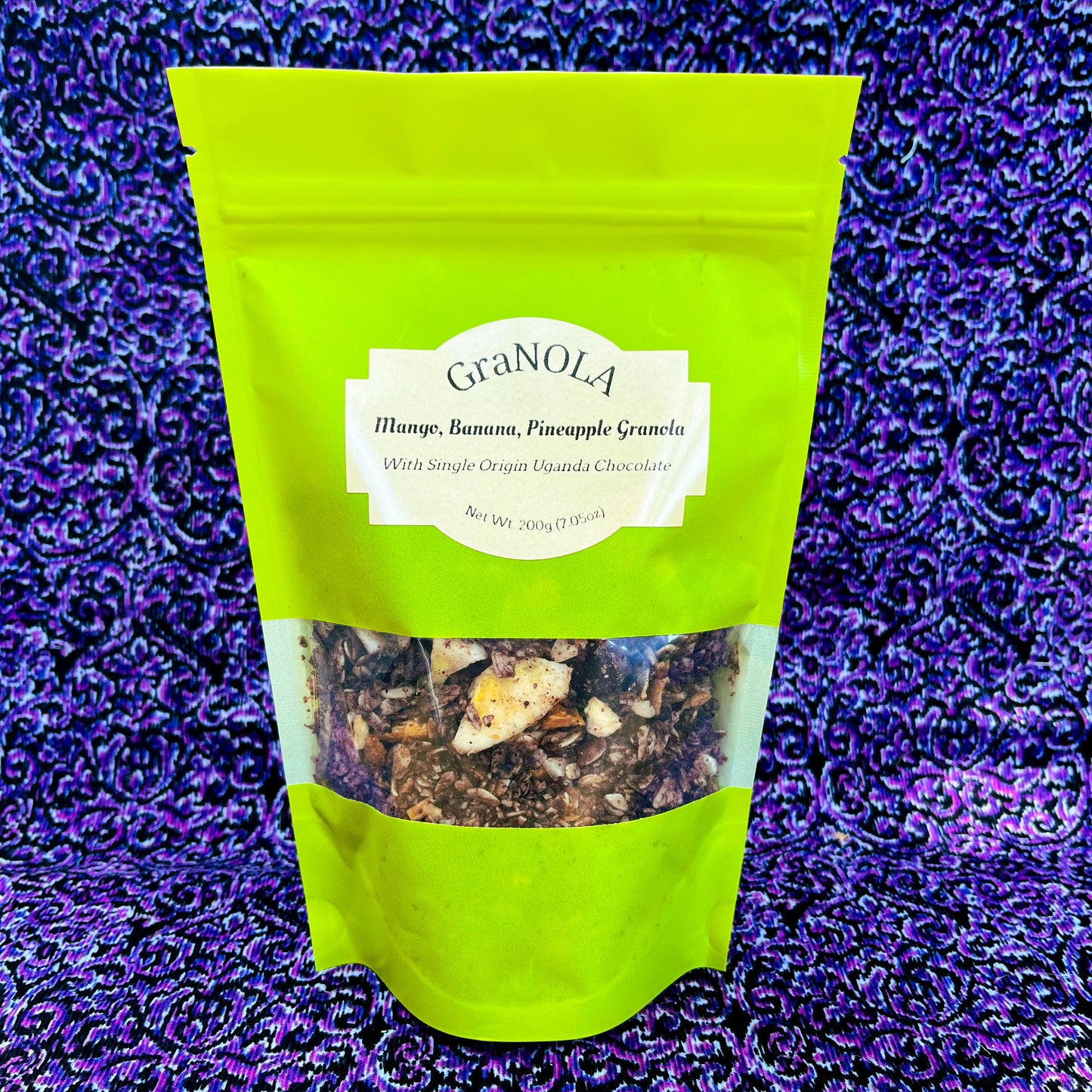 A bright green full of chocolate covered granola with chunks of chocolate, banana chips, mango, and pineapple in it. In front of a purple and black swirled background.