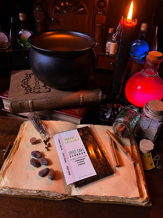 A dark chocolate bar on top of an old notebook surrounded by various spices, potion bottles, a stack of books with a cauldron, and some lit candles.