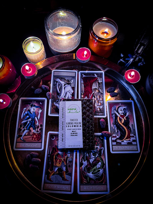 A dark chocolate bar is on top of a round lay out of tarot cards on a gold tray surrounded by lit candles.