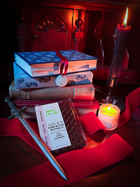 A dark chocolate bar leaning against a stack of books surrounded by candles and red ribbon, with a dagger placed ever so slightly in front.