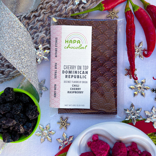 A packaged dark chocolate bar with red chilis, a white dish of freeze dried raspberries, and a green bowl of dried cherries. All surrounded by holiday decor such as red and silver ribbons, green placemat, grey knitwear, and silver snowflake beads.