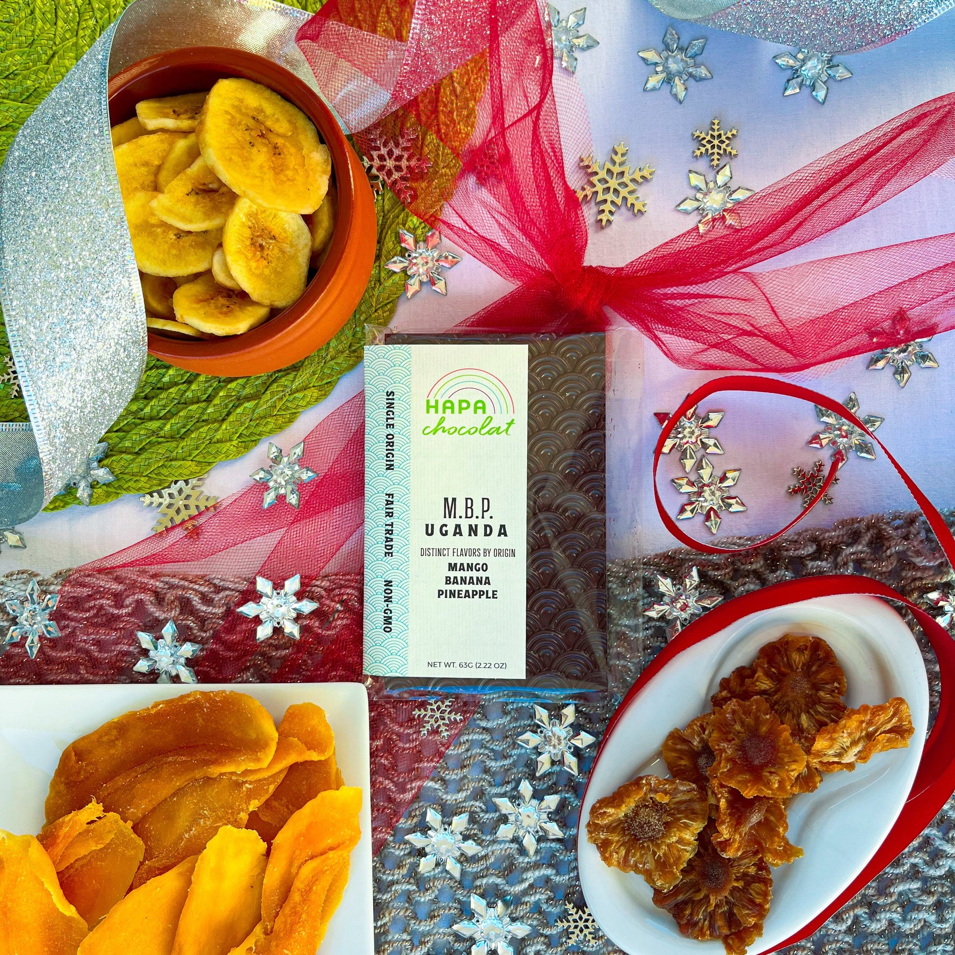 A packaged dark chocolate bar with a white dish of dried mango, a white dish of dried pineapple, and a orange bowl of  banana chips. All surrounded by holiday decor such as red tulle, green placemat, grey knitwear, and silver ribbon and snowflake beads.