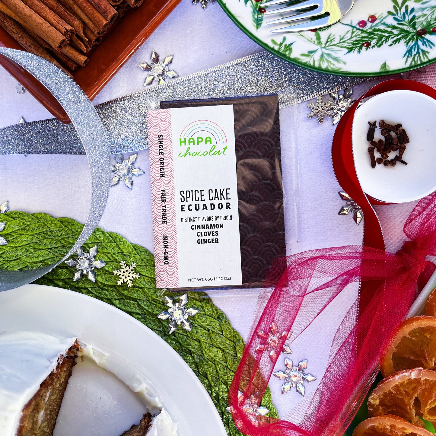 A packaged dark chocolate bar with a whole cake, a slice of cake, a green tray of dried orange slices, a white bowl of whole cloves, and a dish of cinnamon sticks. All surrounded by holiday decor such as. red tulle and ribbon, green placemat, and silver ribbon and snowflake beads.