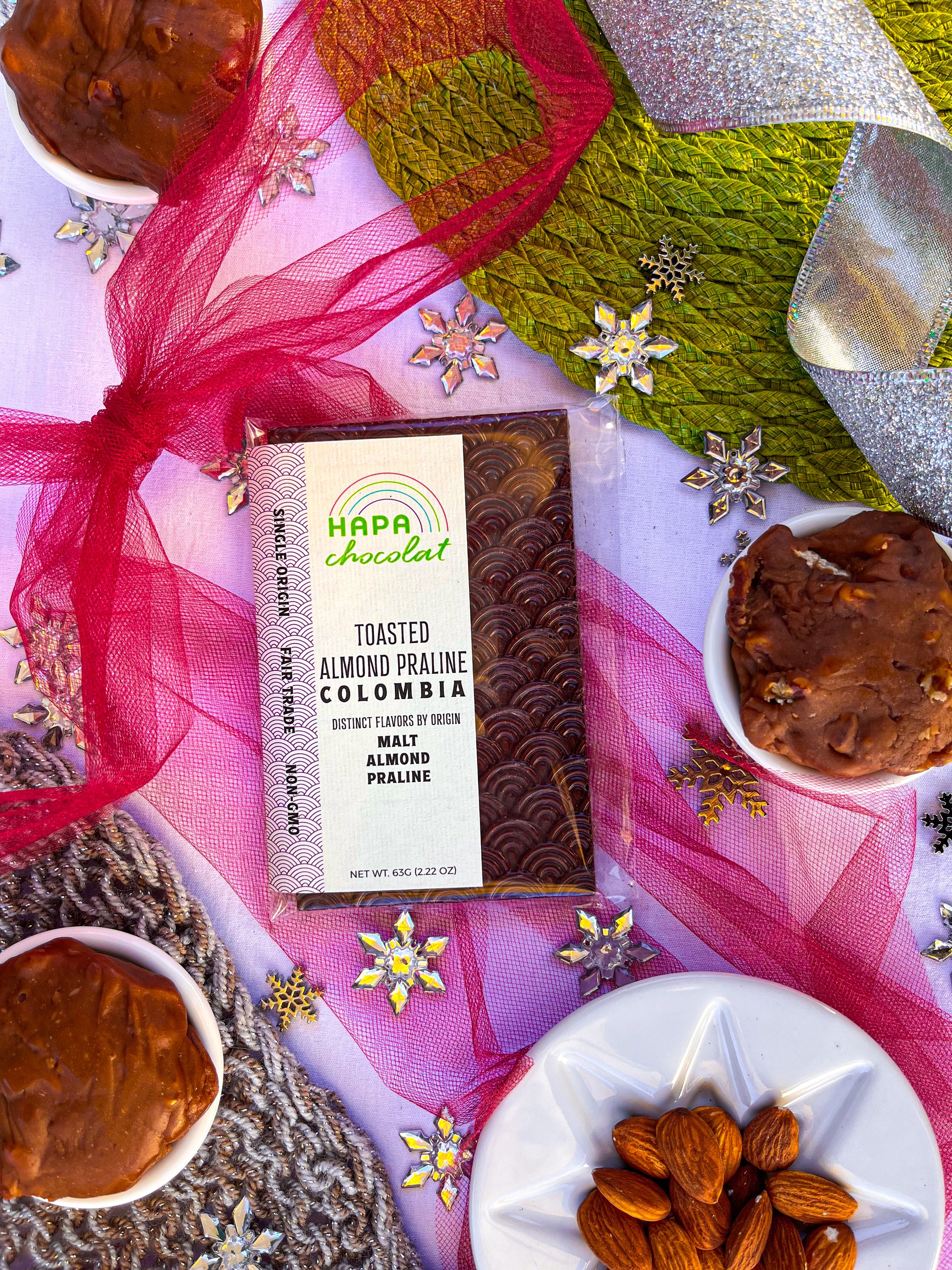 A packaged dark chocolate bar with Southern pralines and a white dish of whole almonds. All surrounded by holiday decorations such as a red tulle bow, silver glitter ribbon, green placemat, grey knitwear, and silver snowflake beads.