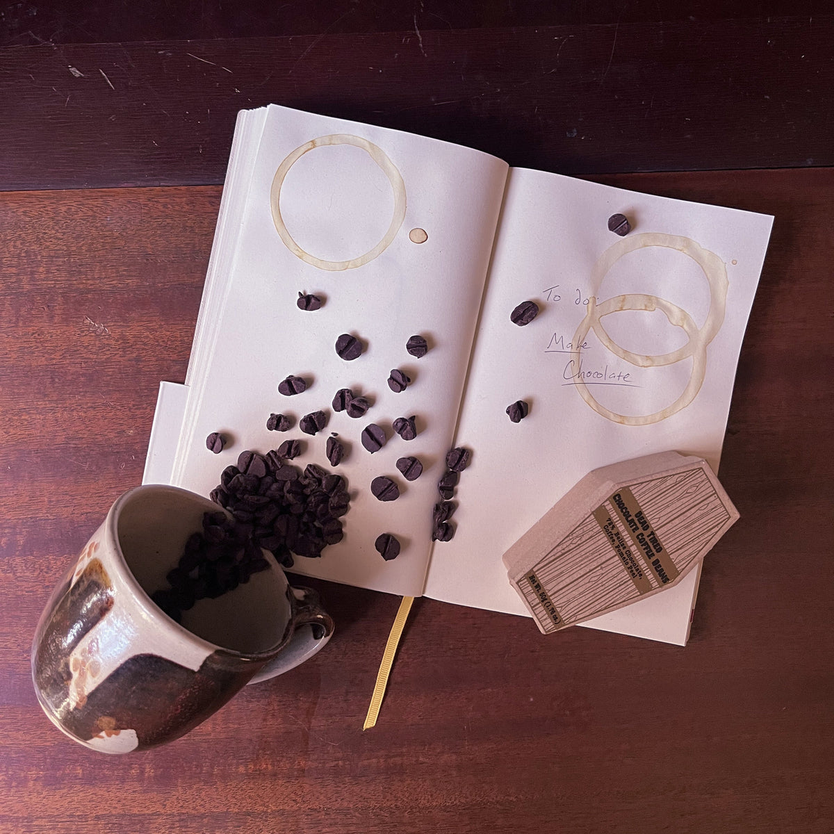A brown and tan ceramic mug is tipped over on a notebook and spilling coffee bean shaped chocolate. The notebook is stained with coffee rings and has 'To Do: Make Chocolate' written on one of the pages. Below the message lays a cardboard coffin with 'Dead Tired Chocolate Coffee Beans' written on top.