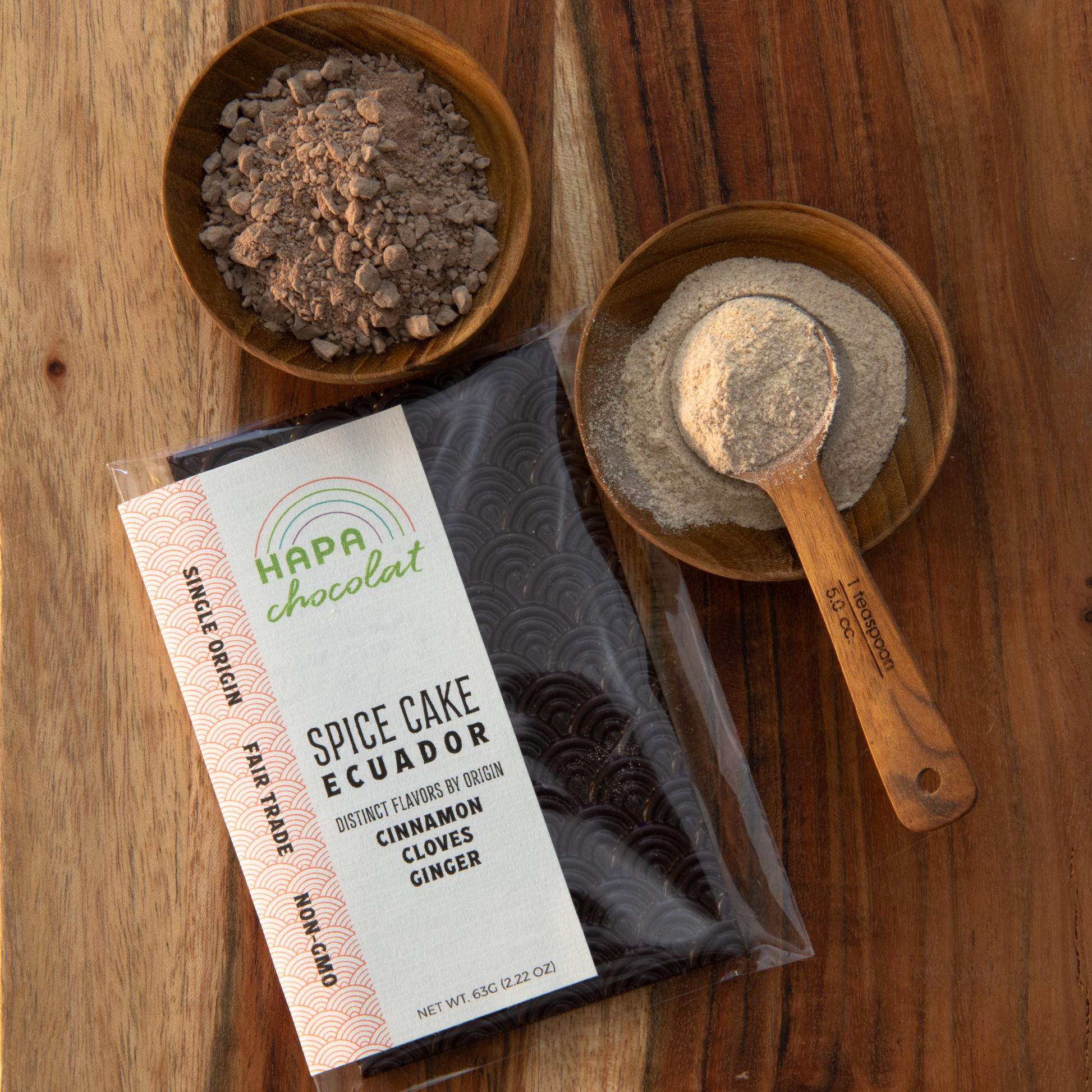 A seigaiha pattern dark chocolate bar in an eco-cello bag with an ivory colored label with the logo and product descriptions. With a wooden bowl of the ground chocolate above it and a wooden bowl of the ground spice brittle and a wooden teaspoon scopping some of the spice up, to the right of the bar.