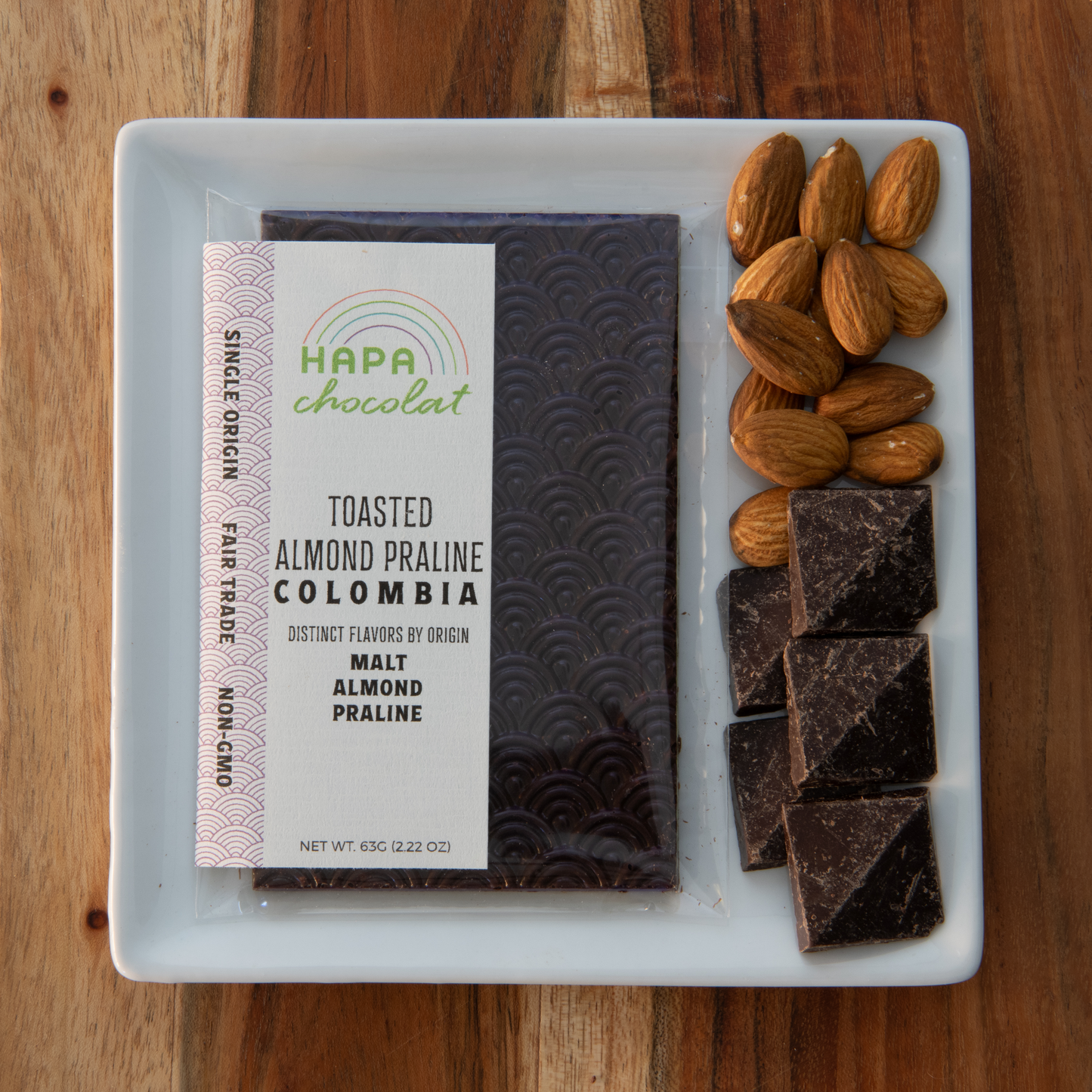 A seigaiha pattern dark chocolate bar in an eco-cello bag with an ivory colored label with the logo and product descriptions one a white plate with whole almonds in the upper right corner of the plate and the large chocolate chips to the left corner or the plate.