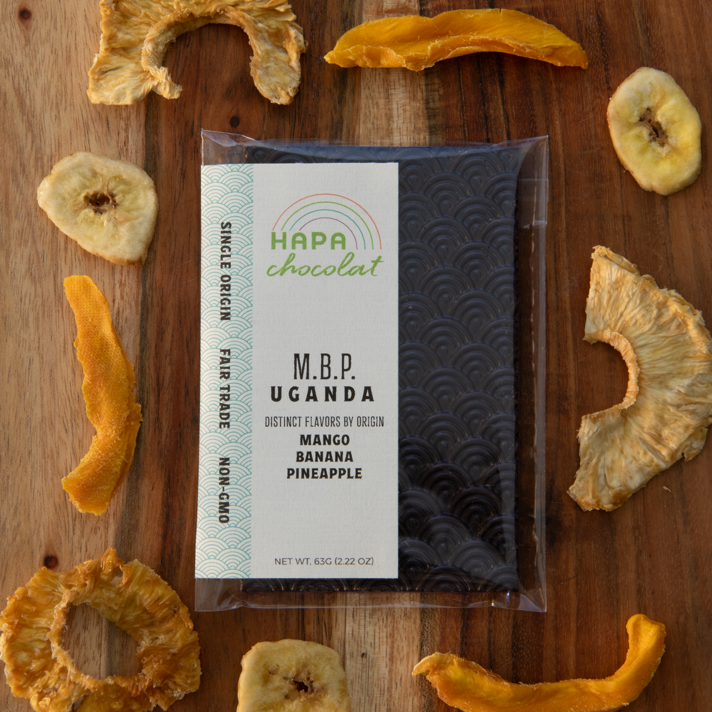  A seigaiha pattern dark chocolate bar in an eco-cello bag with an ivory colored label with the logo and product descriptions, framed by dried mango and dried pineapple pieces, and a few banana chips.