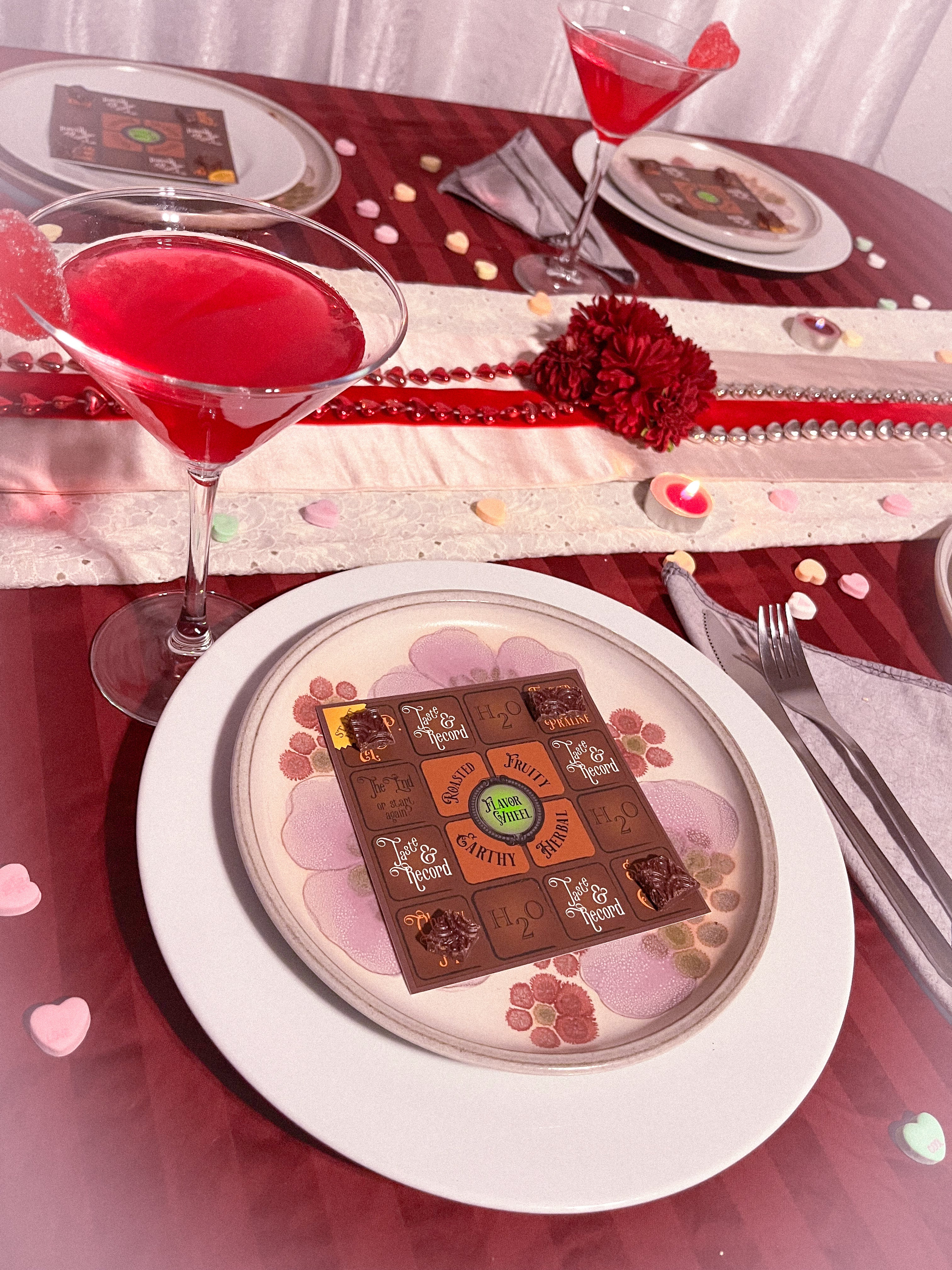 Dinner table set with candles, flowers, and heart candies. There are pink drinks in martini glasses and on each plate is a board game for tasting chocolate.