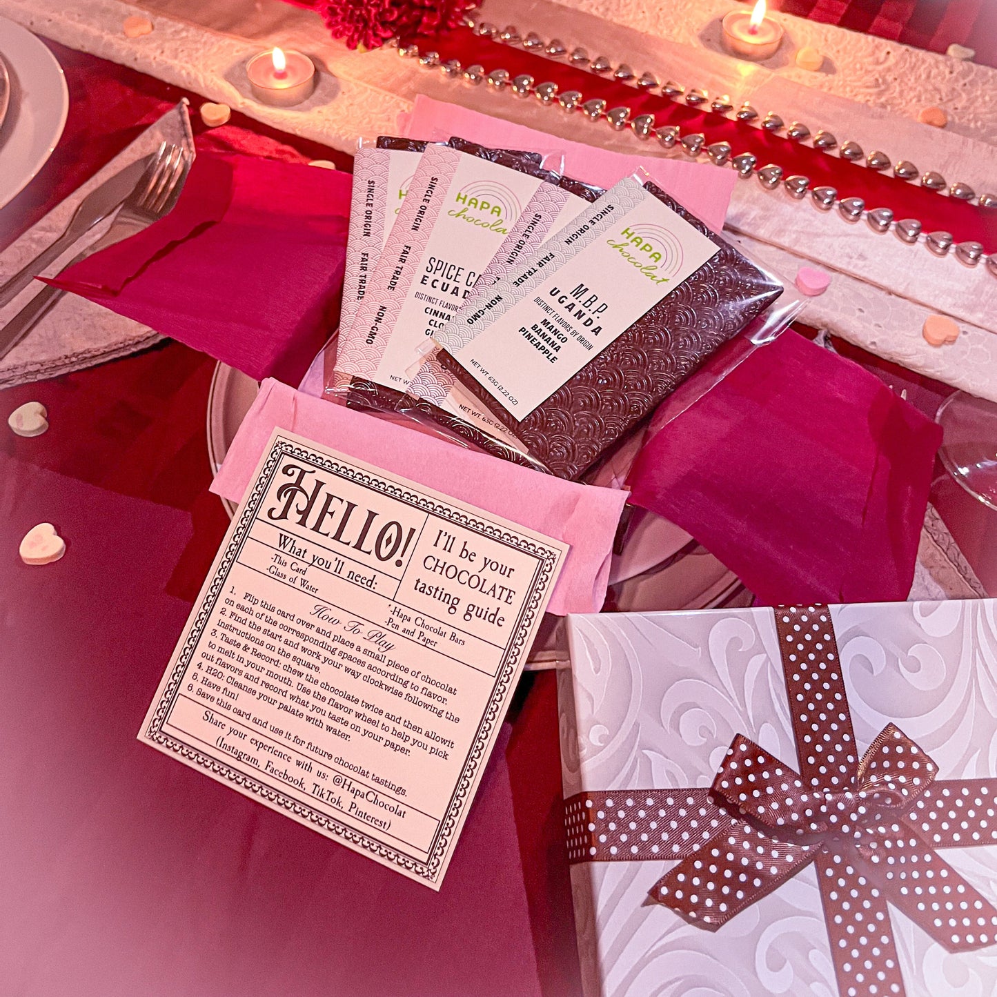 A Valentine's or Galentine's themed dinner is set with candles and flowers and beads. Each place setting has a gift box and one is open to display 4 chocolate bars and an instruction card.